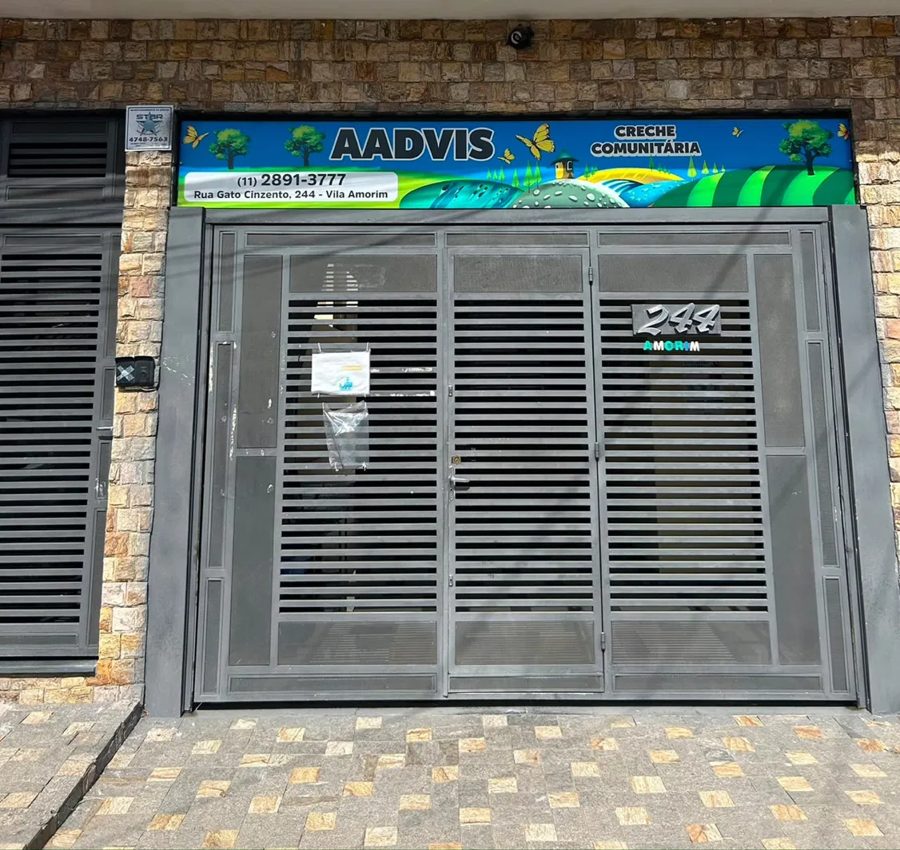 Fachada da Creche AADVIS Vila Amorim, com portão de grade cinza e faixa azul de identificação