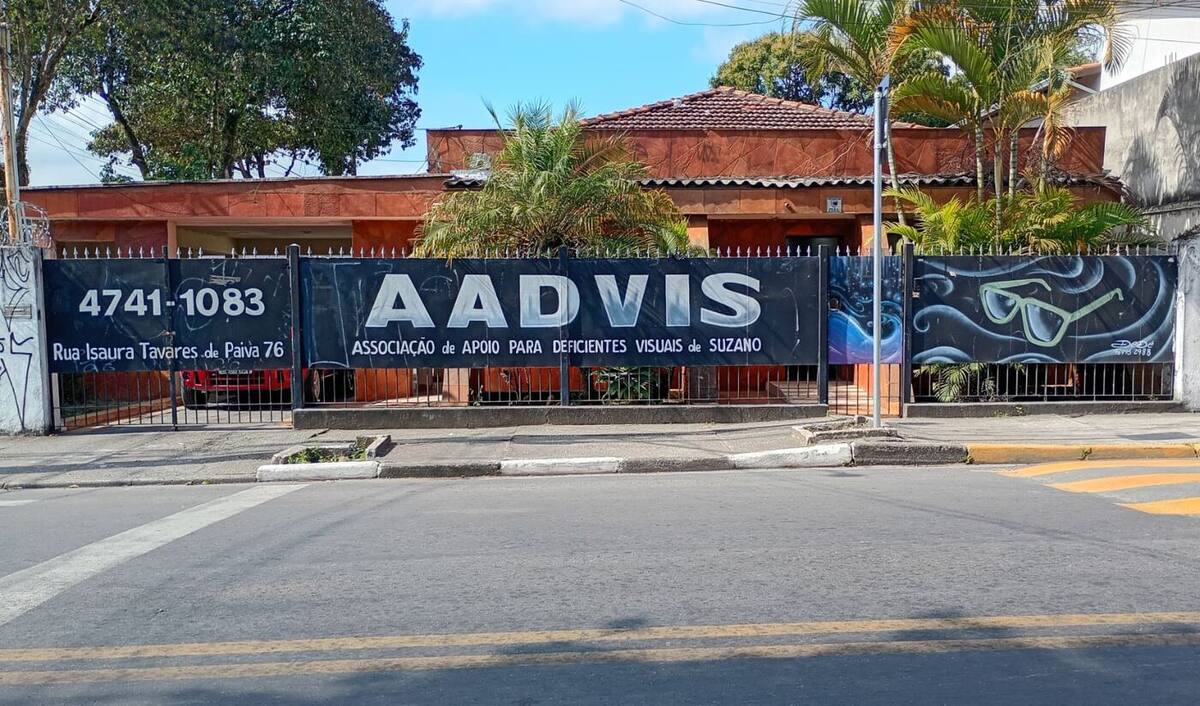 Fachada da sede da AADVIS, uma casa térrea com telhas de barro e área externa arborizada, cercada por um muro com o nome da associação em letras brancas sobre fundo escuro